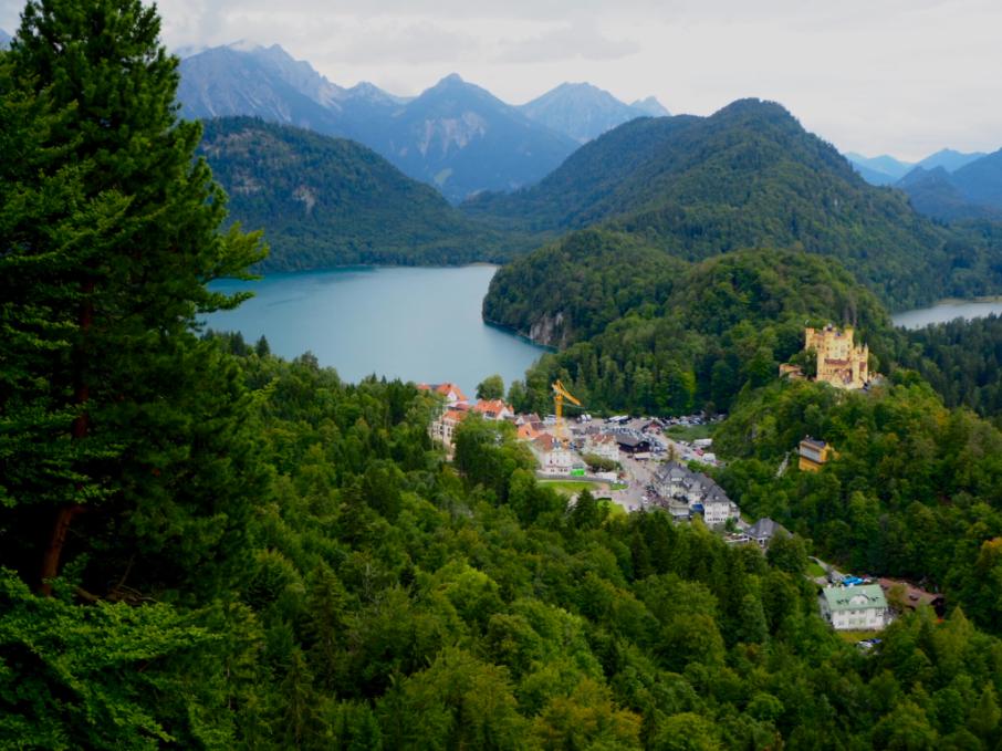 A lovely mountain scene of pine covered foothills, an electric blue alpine lake stands before sharp mountain peaks. A small village along with a yellow castle stand in the bottom right corner below the viewer.