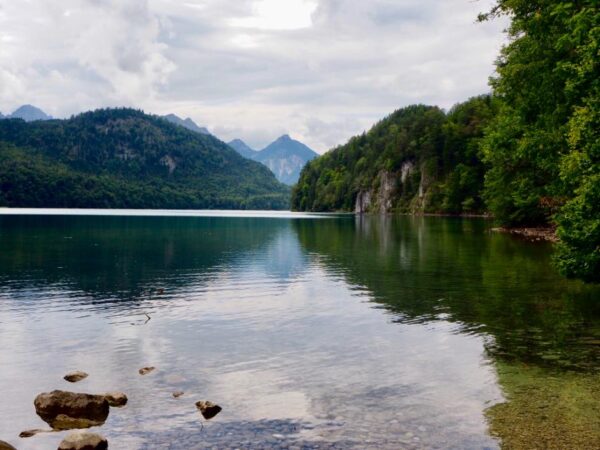 A large lake is shown with a cluster of rocks in the foreground, beneath a cloudy sky. The surface shows only a few gentle ripples and reflects many of the mountain peaks behind it.