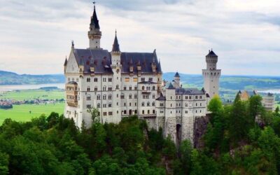A beautiful castle constructed on top of a rocky outcrop stands tall against the backdrop of green plains, lakes and hills behind it. Surrounded by pines and green foilage, multiple spires and turrets top the castle, along with many windows.