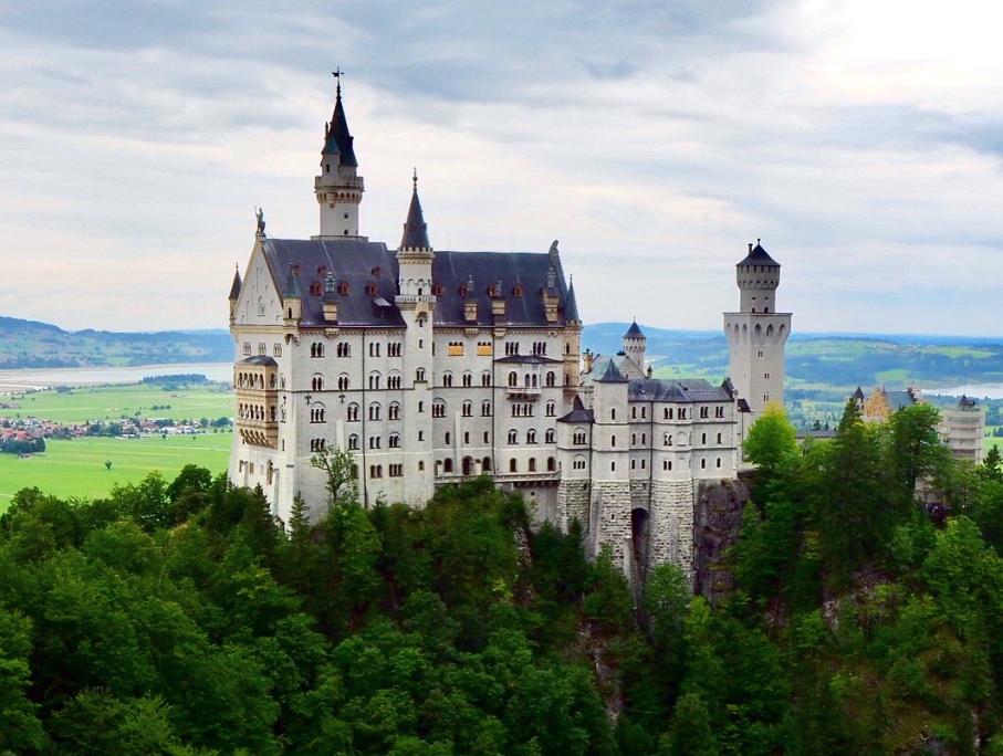 A beautiful castle constructed on top of a rocky outcrop stands tall against the backdrop of green plains, lakes and hills behind it. Surrounded by pines and green foilage, multiple spires and turrets top the castle, along with many windows.